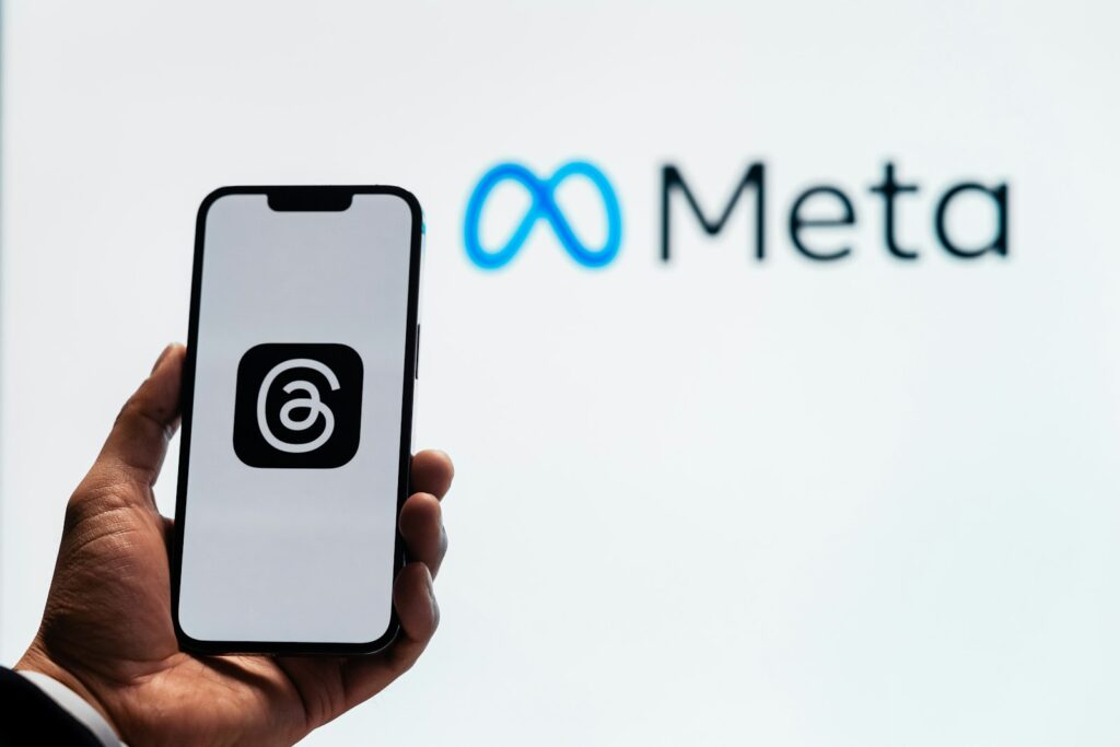 Mao segurando um celular com o app Threads aberto e o logo da Meta ao fundo, ilustrando a ampliacao dos Live Chats no Threads