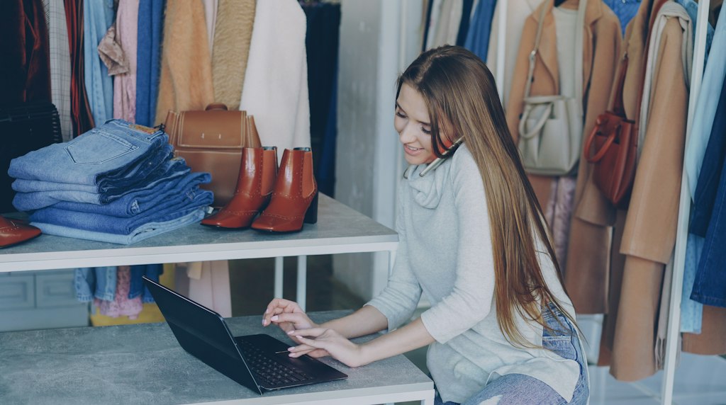 Lojista acompanhando os pedidos da sua loja em um laptop, cercada por produtos